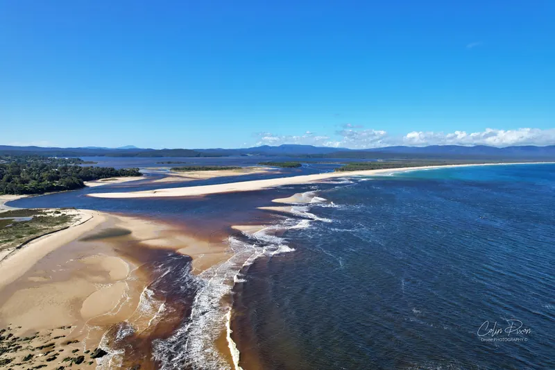 Aerial photo of the Mallacoota Inlet mouth at low tide, with sandbars, surf and dark water.