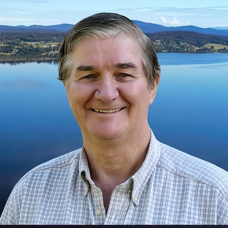 Colin Dixon smiling, in a checked shirt, with Mallacoota Inlet and hills behind him.