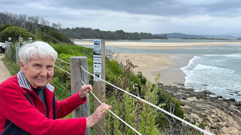 Colin's mum smiling in a red jacket at the lookout fence at Bastion Point, with the beach and ocean behind her.