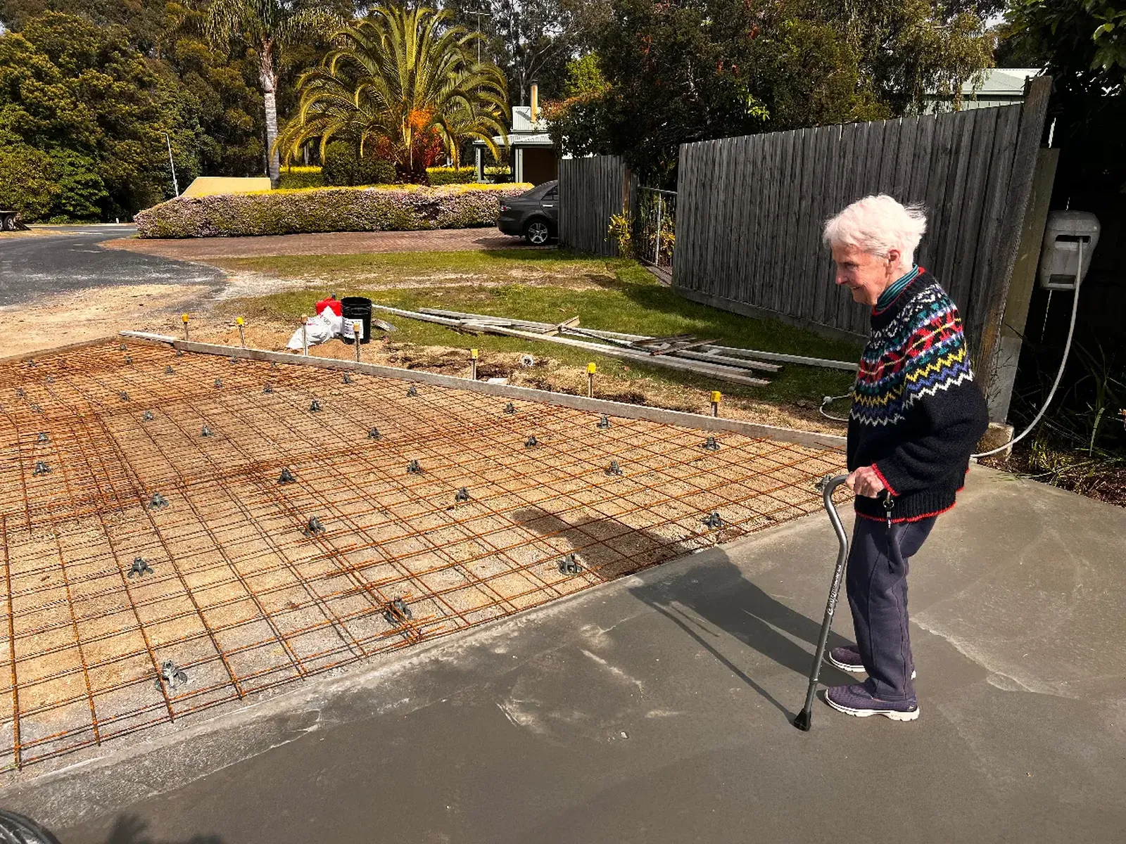 An elderly woman with a walking stick on a new concrete path, looking at the steel reinforcing mesh laid for the driveway.