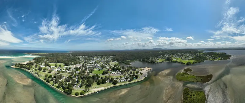 Drone panorama of Mallacoota township on its peninsula in the inlet, with sandbanks, bush and blue sky.