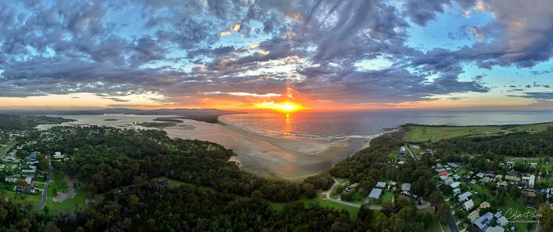 Drone panorama of sunrise over Mallacoota Inlet, with houses and bush in the foreground and cloud lit orange.